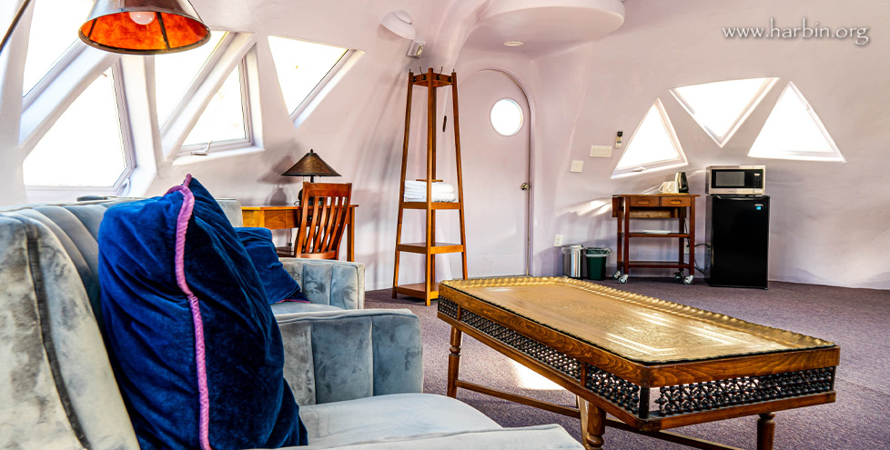 The inside of a dome shaped Dome room has colorful purple and blue decor with a coffee table, large lounge chairs, a microwave and small fridge sit in the background. The triangle shaped windows let light into the bright room.