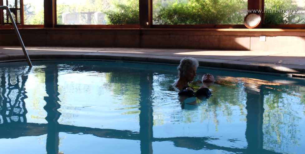 A person receives a health treatment in the therapy pool, being floated by the silouette of the practitioner. In the background windows let in light and plants are outside. 