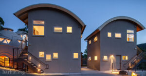Two unique two story structures with windows lit in the evening sky have curved roofs. Beside stands a round smaller building. 